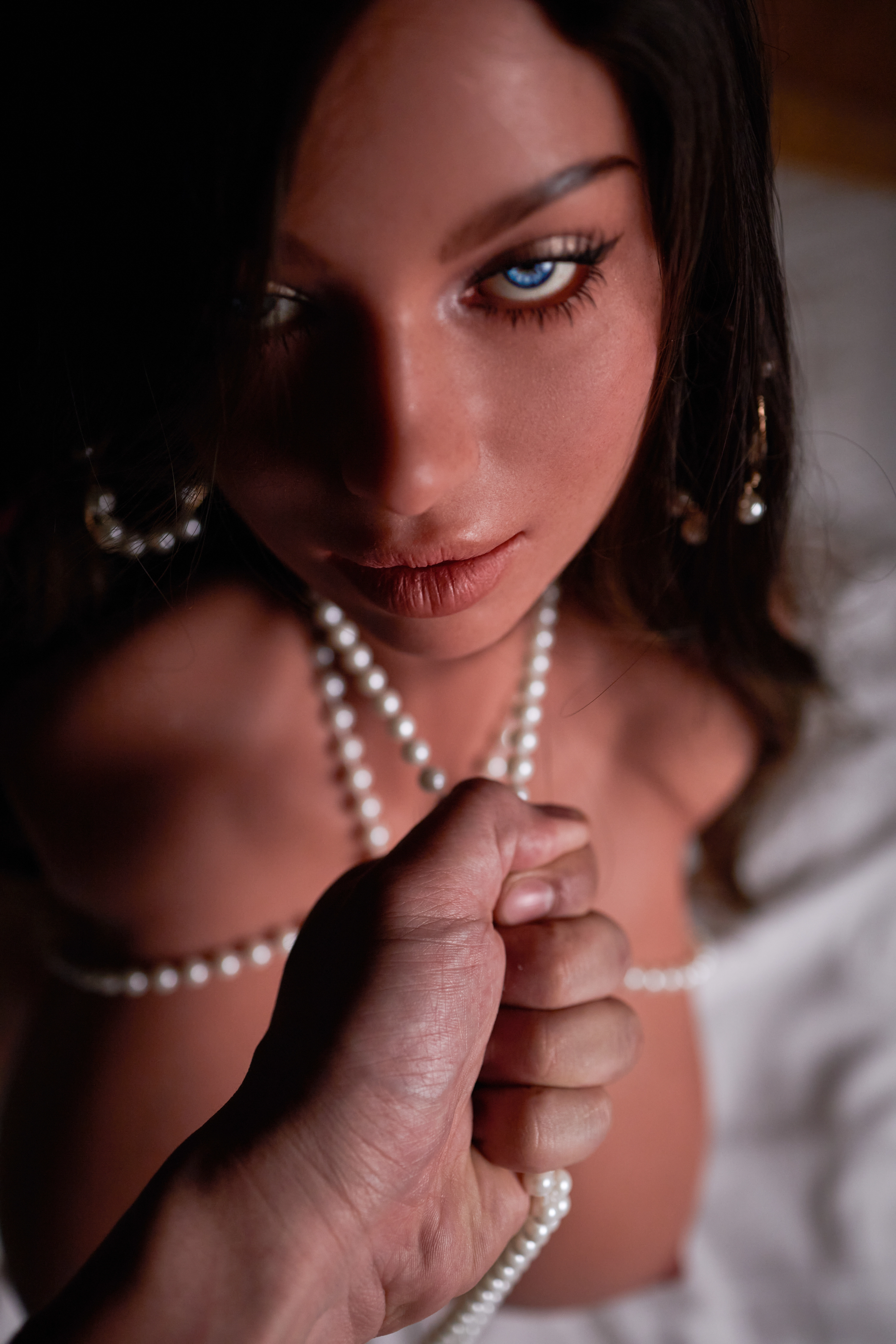Close-up portrait of a woman with long dark hair, striking blue eyes, and soft makeup, adorned with pearl necklaces and earrings, with a hand holding a strand of pearls.